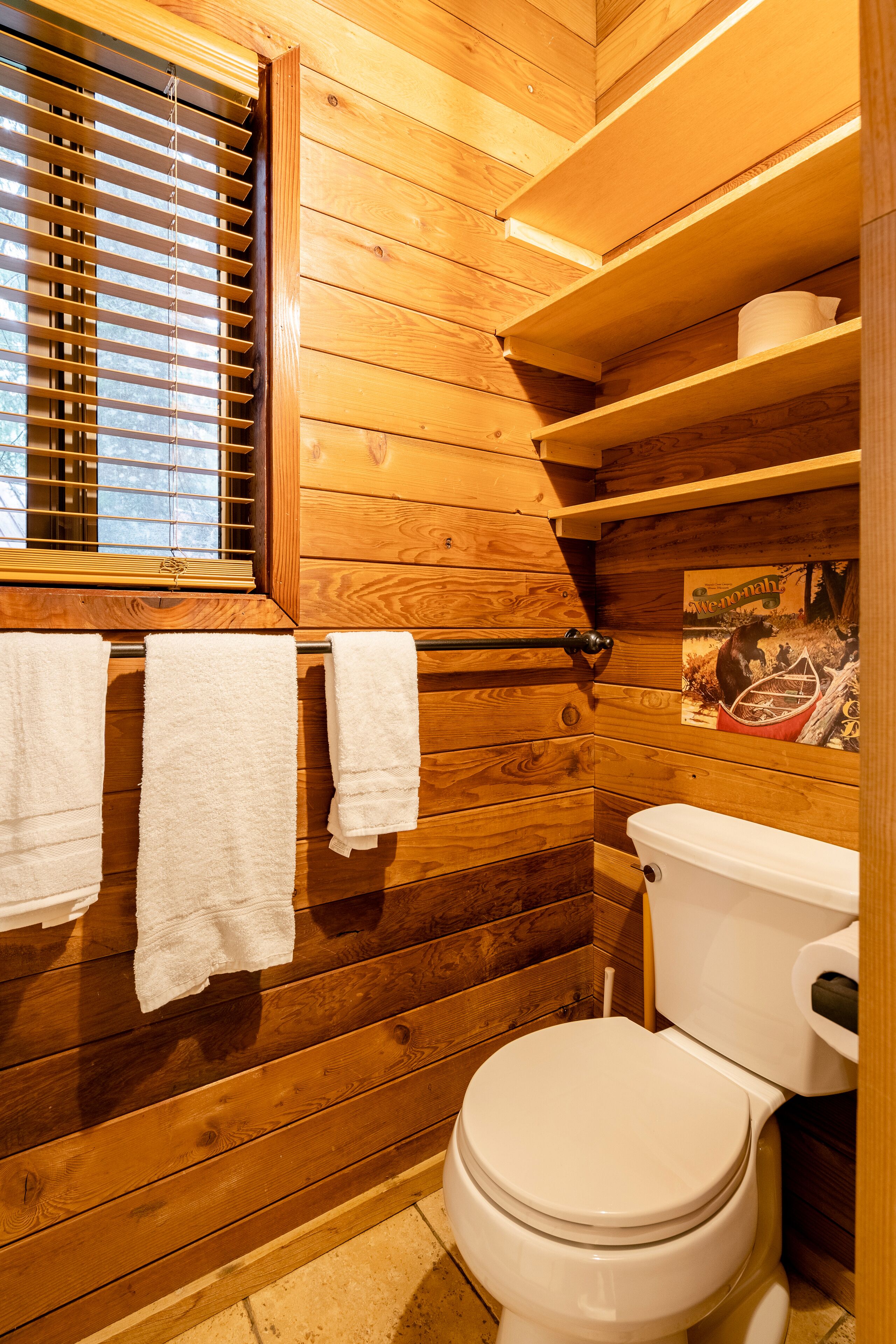 Een badkamer in chaletstijl met houten lambrisering, een toilet, houten planken en een raam met zonwering.