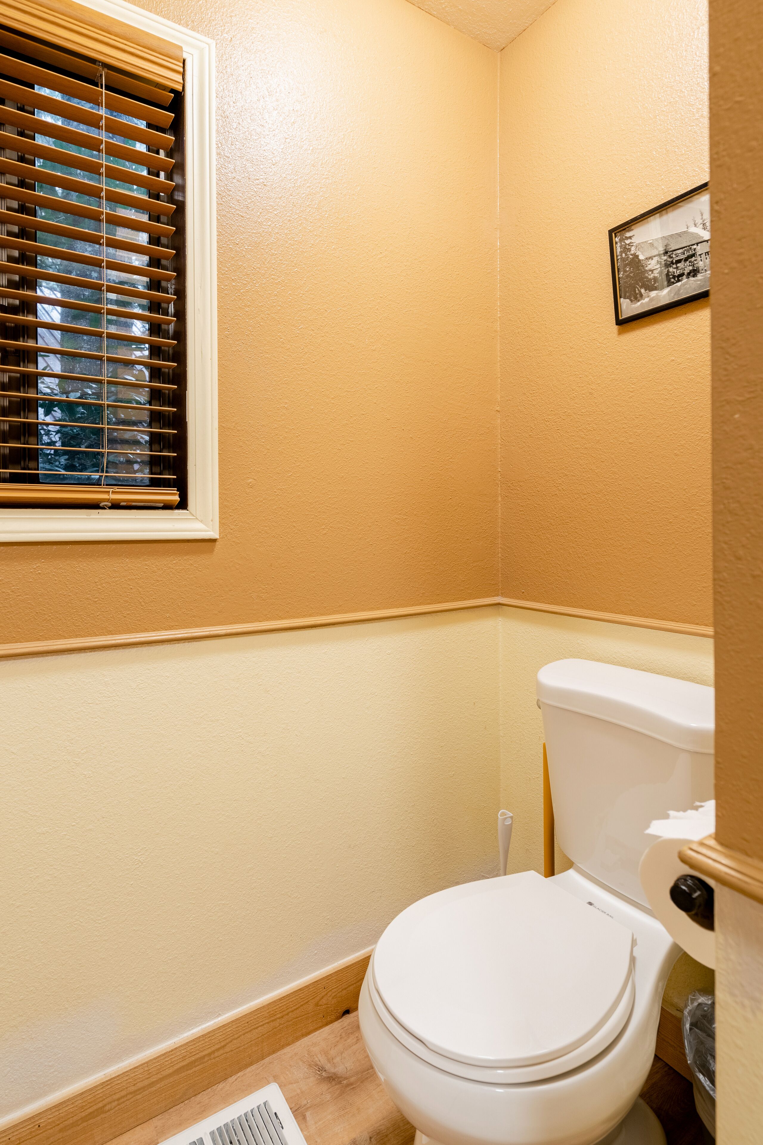 A small, well-lit bathroom with a toilet, a window with blinds, and a framed picture on the wall.