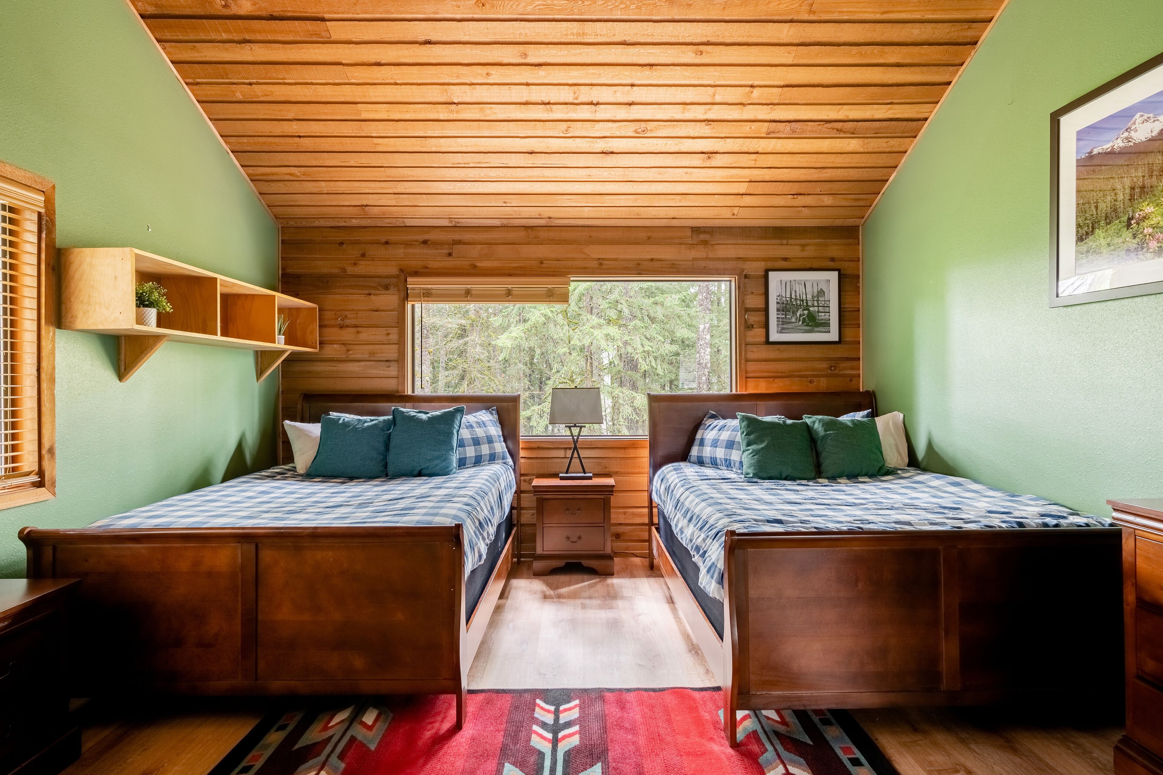 A cabin-style bedroom with two wooden beds, a nightstand, a shelf with a plant, and a window with a view of trees.