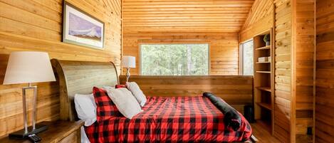 A wooden cabin bedroom with a bed, bedside lamps, a nightstand, a window, and a shelf with a plant.