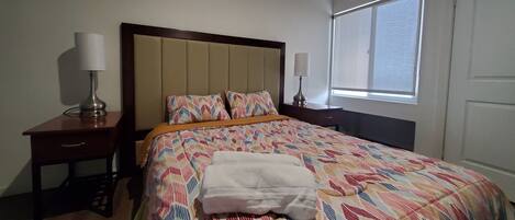 Una cama bien hecha con una colcha estampada, dos lámparas de noche, una mesita de noche de madera con un cajón y una ventana con persianas.