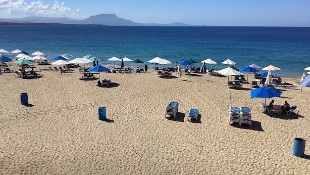 Une plage bordée de rangées de chaises longues et de parasols, un ciel dégagé et une montagne qui se profile à l’horizon.