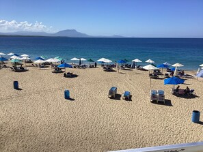 Una playa con hileras de tumbonas y sombrillas, cielo despejado y una montaña a lo lejos.
