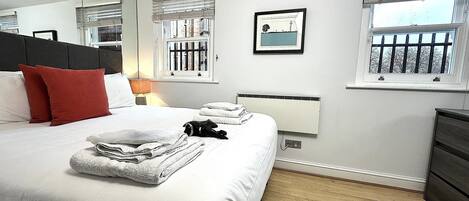 A neatly made bed with white linens and red pillows, a bedside lamp, and a window with blinds.
