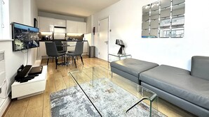 Un soggiorno moderno con un divano grigio, un tavolino da caffè e una TV montata a parete. Si intravede in secondo piano una cucina con mobili bianchi e un tavolo da pranzo.