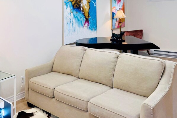 A beige sofa in a modern living room with a glass coffee table, abstract paintings, and a small table with a lamp. - Basement Guesthouse near National Park and Montreal (St-Bruno-de-Montarville)
