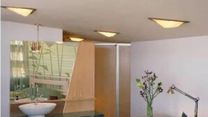 Et moderne værelse med skrivebord med glasplade, en bærbar computer, en skrivebordslampe, en stol, et lille bord med en vase blomster og et stort spejl.