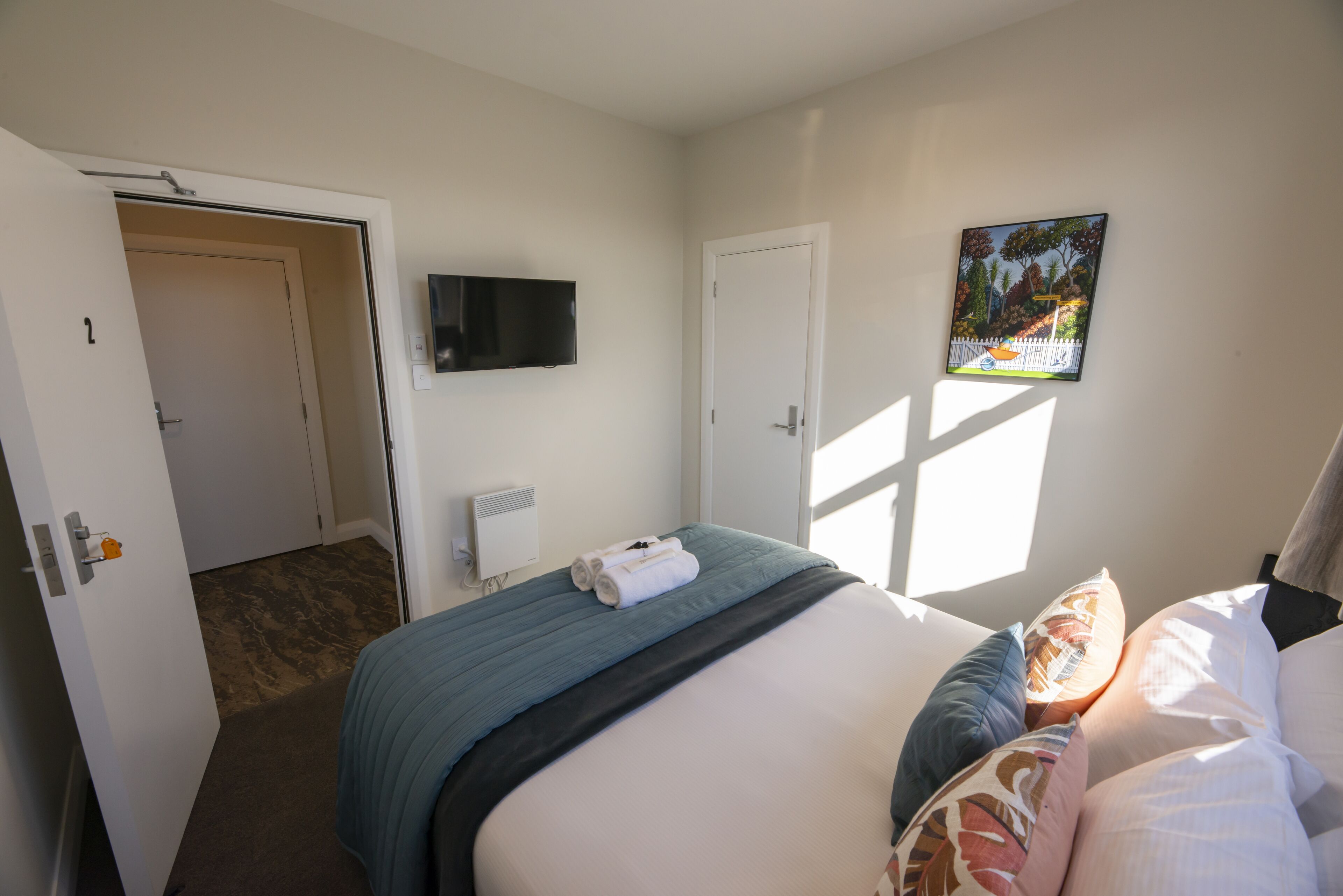 A hotel room with a bed, a television, a painting, and a door leading to another room.