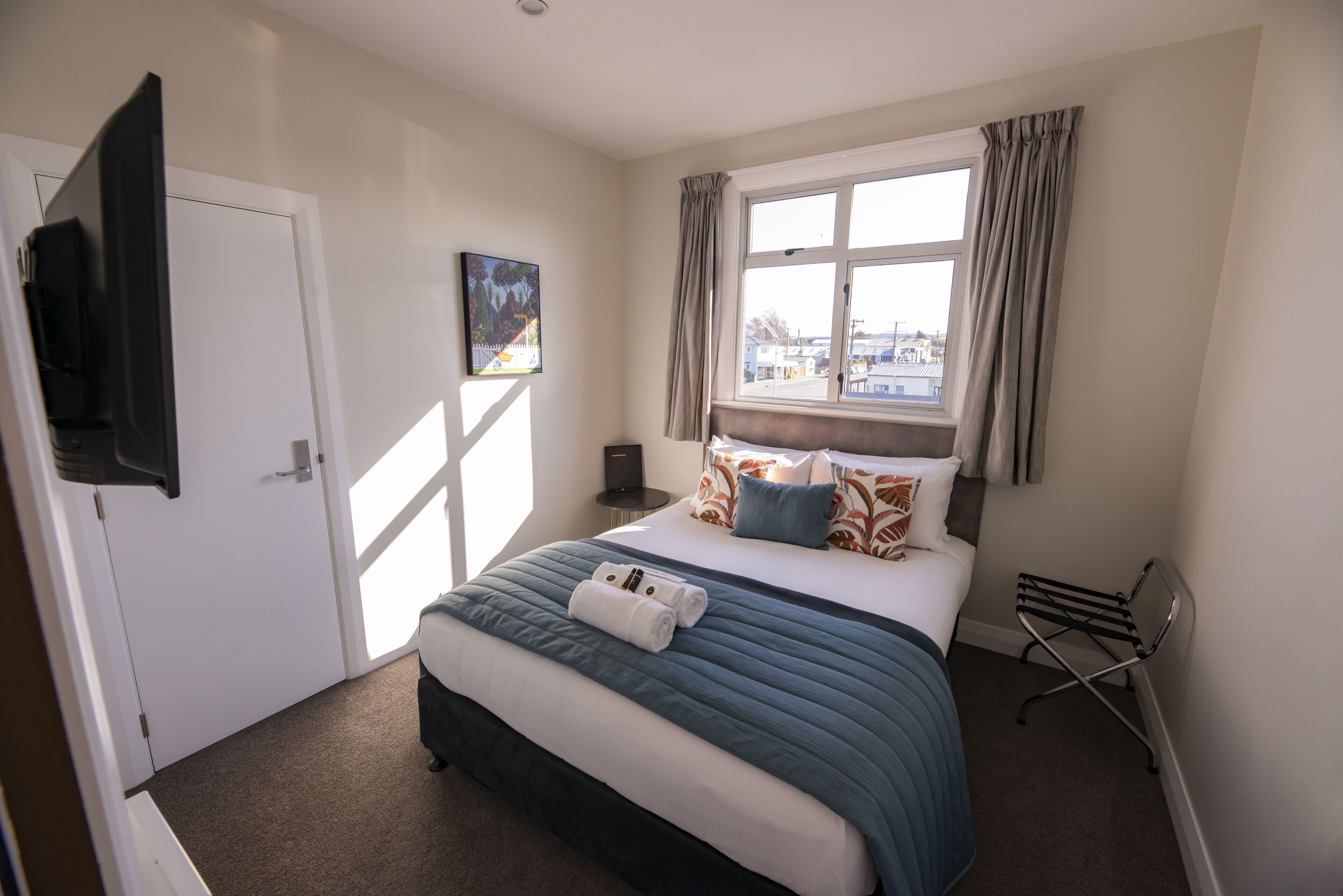 A hotel room with a bed, a flat-screen TV mounted on the wall, a window with curtains, and a painting on the wall.
