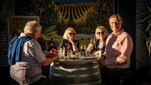 Un grupo de personas sentadas alrededor de una mesa de madera con copas de vino, en un entorno al aire libre con un viñedo al fondo.