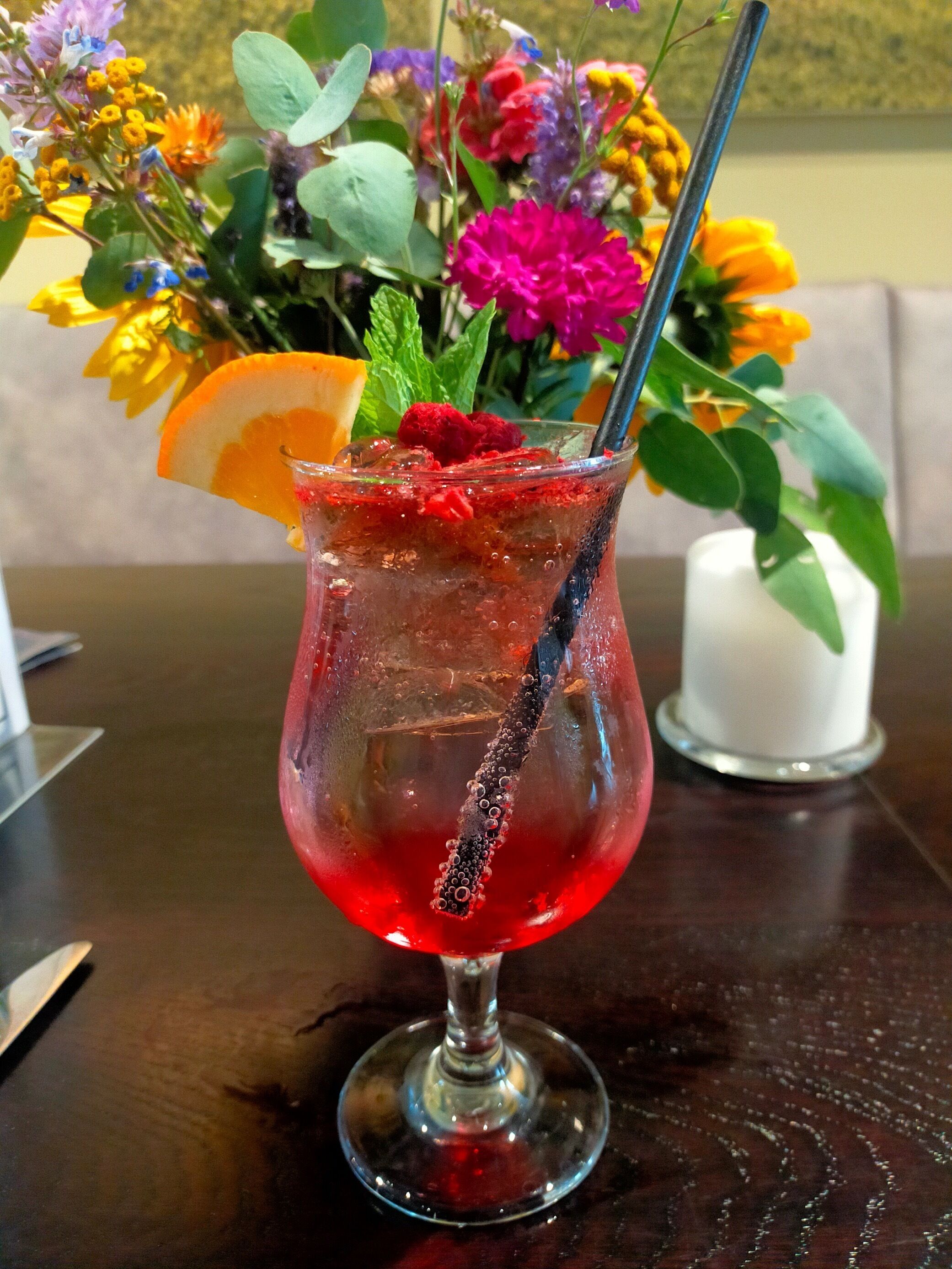 Um cocktail com guarnição de fruta e canudo, sobre uma mesa com um arranjo floral ao fundo.