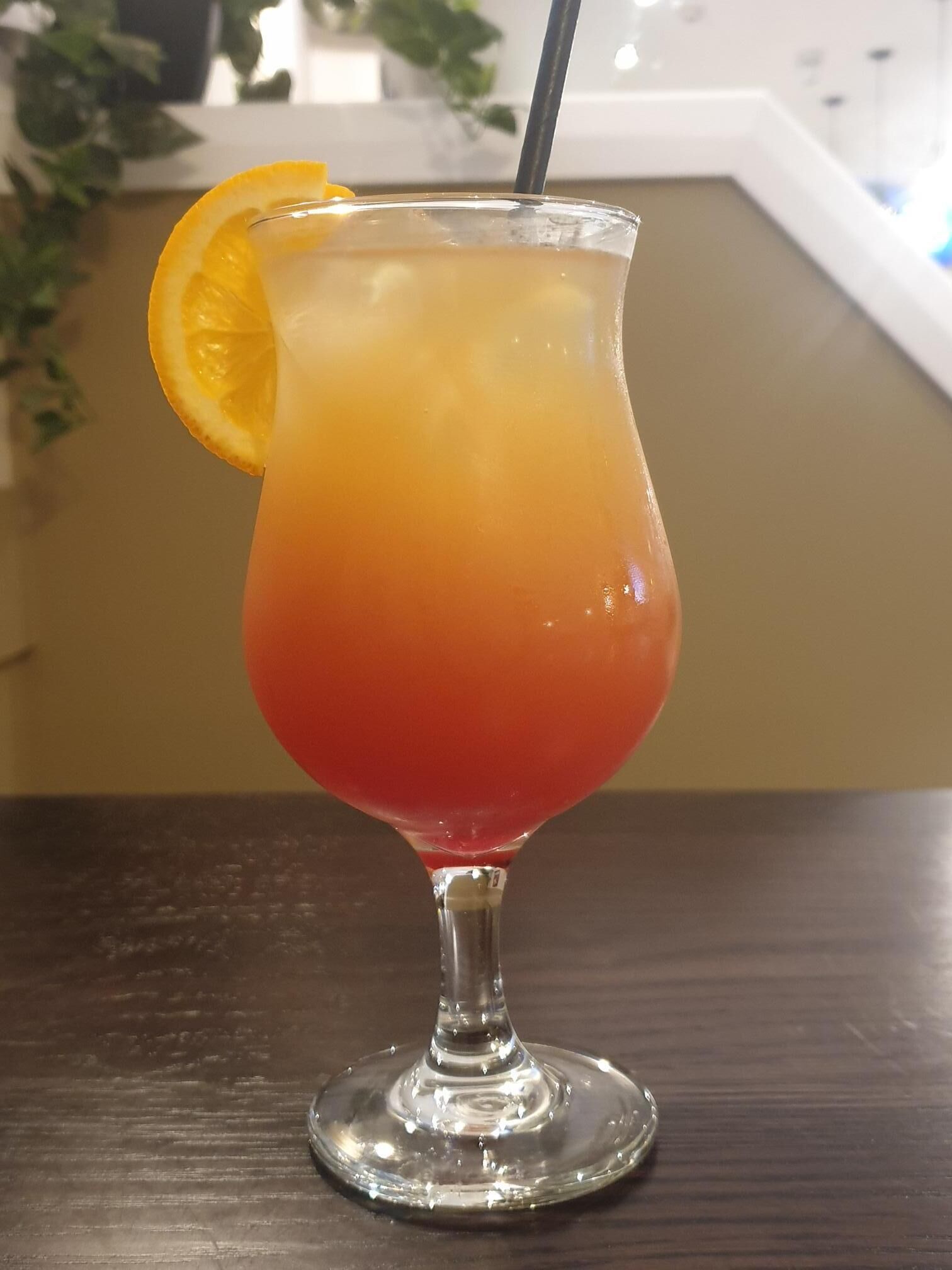 En cocktail i et glas med farvegradient fra rød til gul, pyntet med et appelsinstykke og et sugerør.
