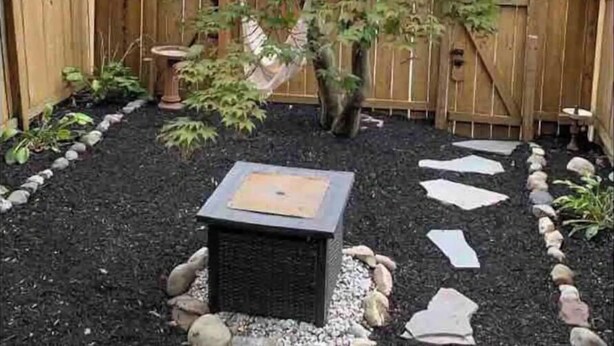 Un jardín con cerca de madera, un árbol, piedras para pisar y una jardinera cuadrada.