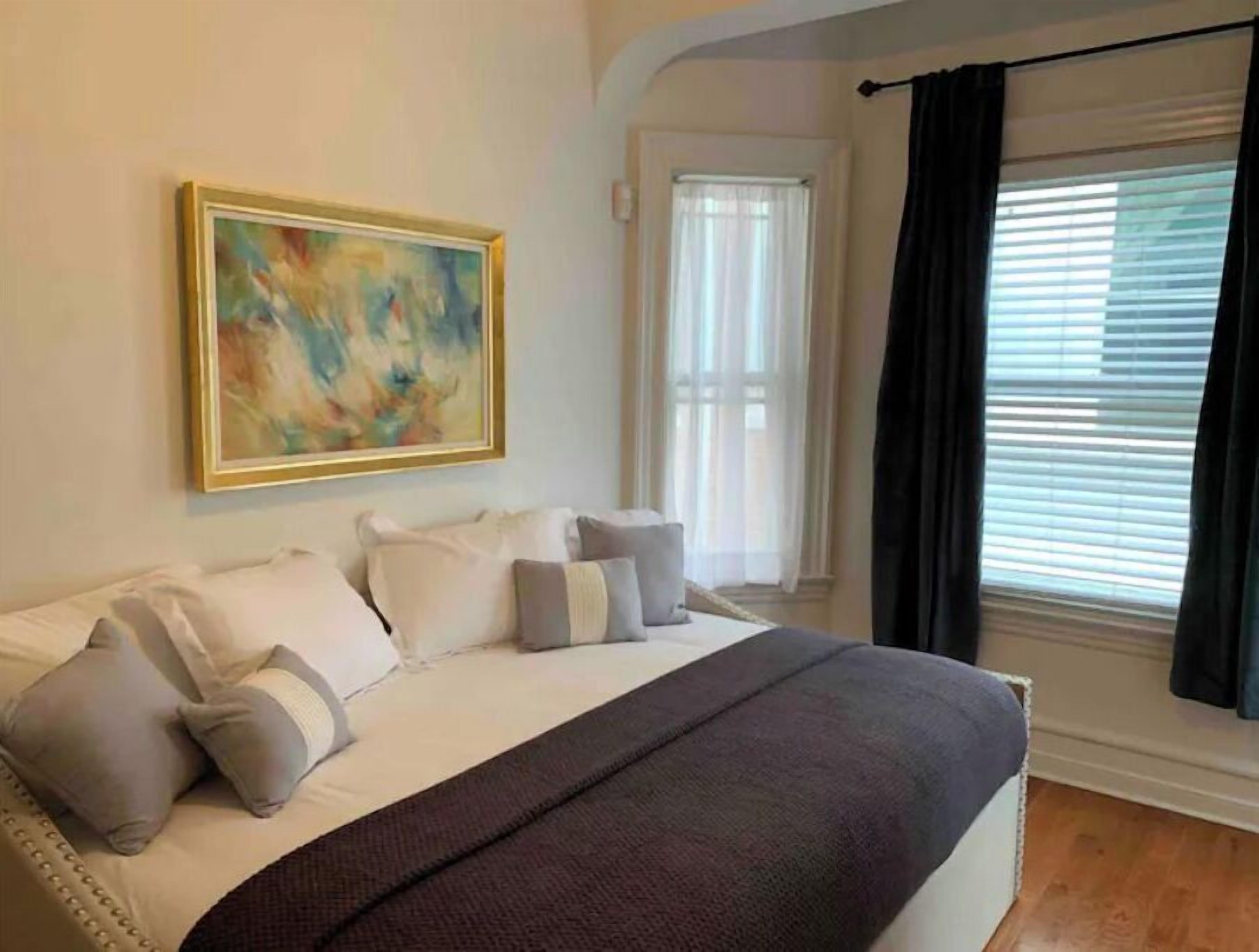 Une chambre à coucher avec un lit, des oreillers, une couverture, un tableau et une fenêtre avec des rideaux.