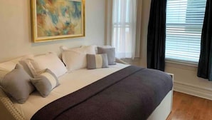 Un dormitorio con cama, almohadas, una colcha, un cuadro y una ventana con cortinas.