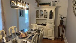Comedor con una mesa de madera oscura dispuesta para cuatro personas, sillas blancas, lámpara colgante y un armario blanco con puertas de vidrio.