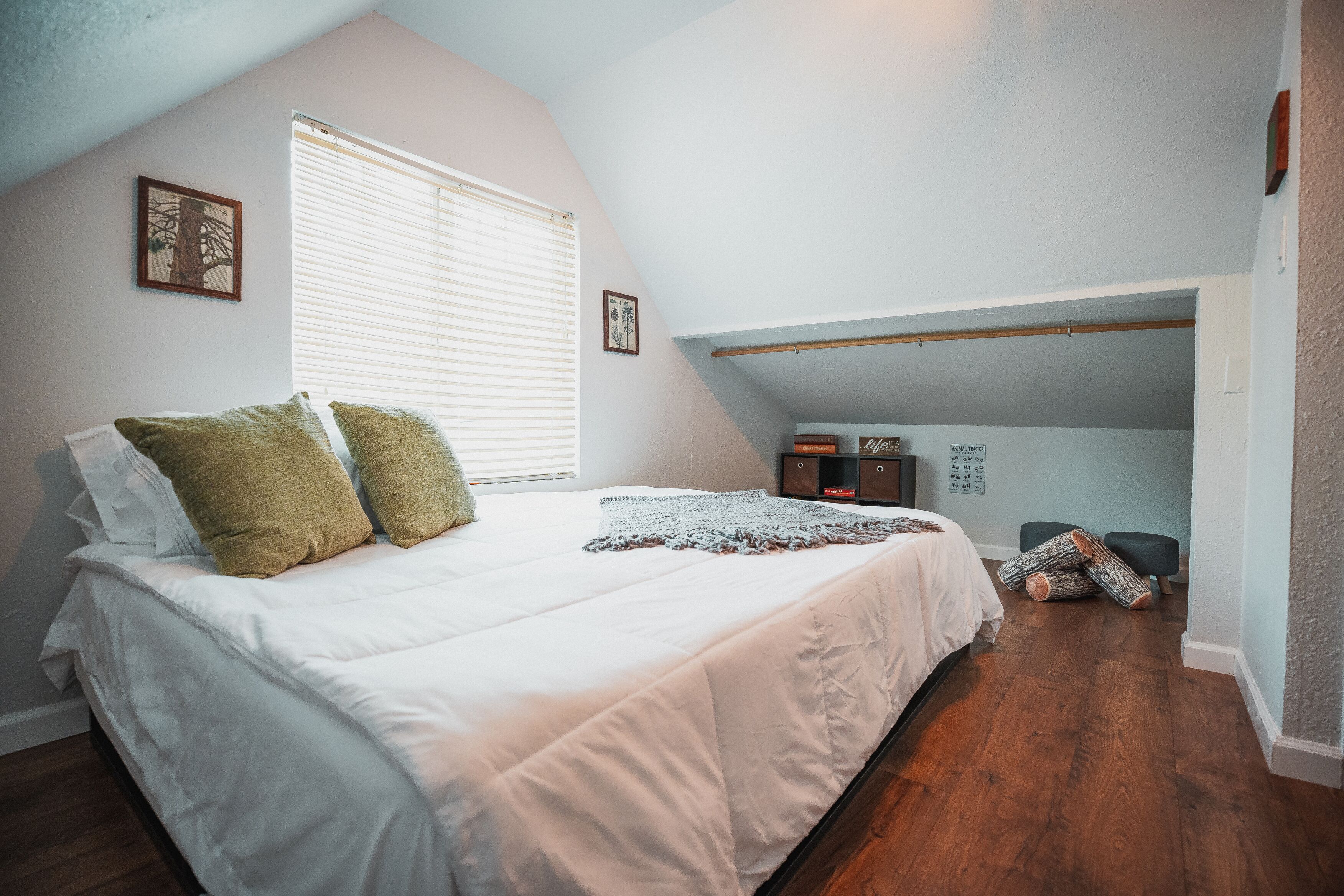 A bedroom with a sloped ceiling, a bed with white bedding and green pillows, a window with blinds, and a small desk with a chair.