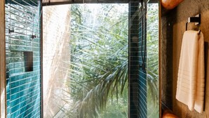Een badkamer met een grote glazen deur naar uitzicht op een tropische tuin, betegelde muren en een houten bankje met een handdoek.
