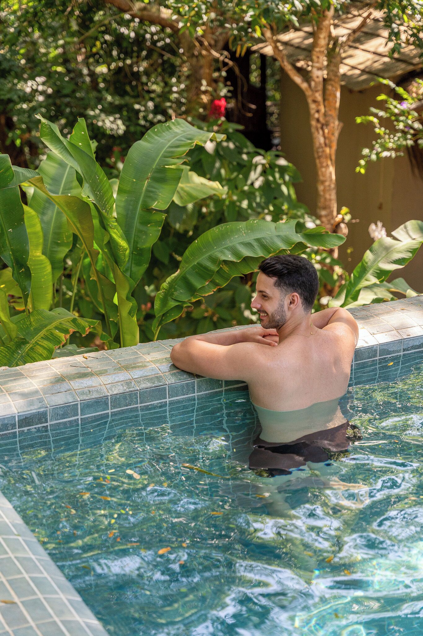 A man relaxing in a swimming pool with tiled edges, surrounded by lush greenery and a tree.