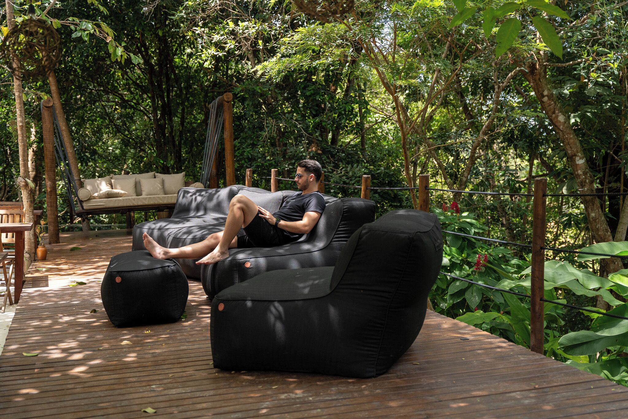 Une personne se détend sur un canapé noir installé à l’extérieur, agrémenté d’un coussin et d’un pouf, et est entourée d’une végétation luxuriante.