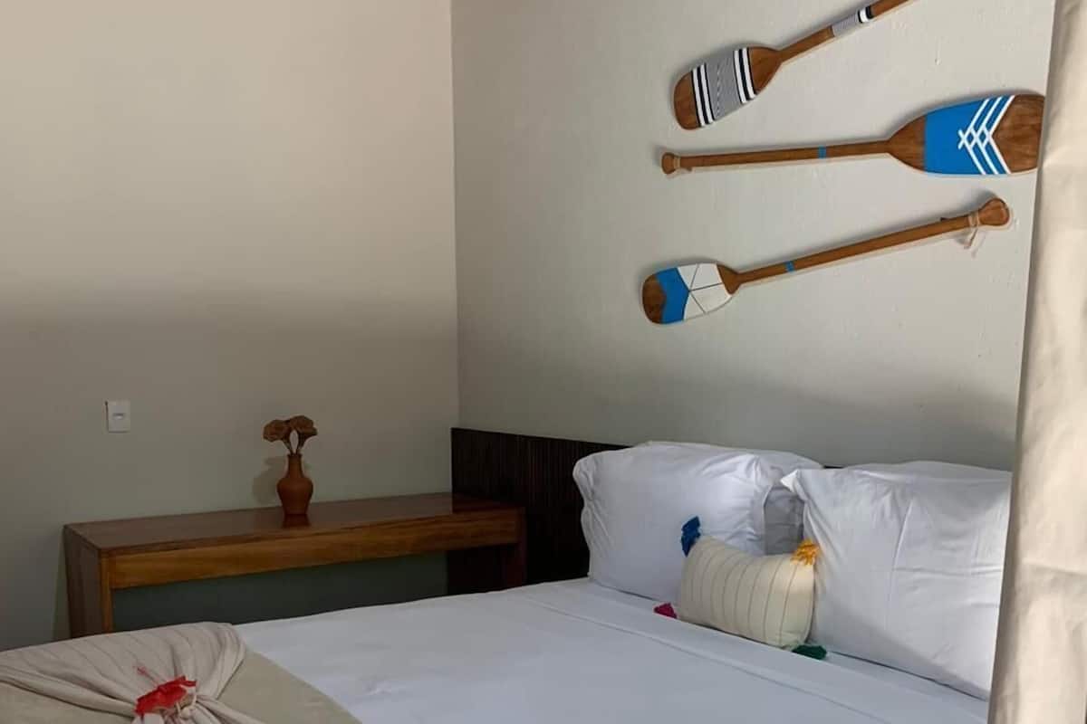 Een slaapkamer met een bed, kussens, een nachtkastje, een vaas en twee houten scheen op de muur.