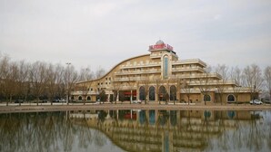 Un grand bâtiment à plusieurs étages, doté d’une entrée centrale en forme d’arche, qui se reflète dans une étendue d’eau, et dont le toit plat est surmonté d’une enseigne rouge.