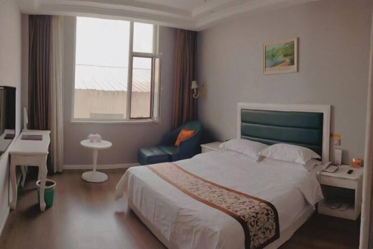 A hotel room with a bed, a desk, a chair, a small table, a window, and a painting on the wall.