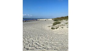 Una playa arenosa con cielo azul despejado, algo de vegetación a la derecha y unas pocas sombrillas de playa a lo lejos.