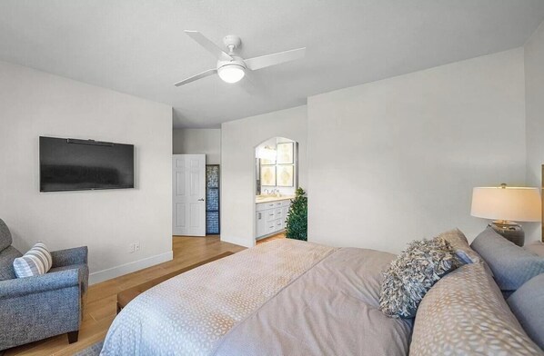 A bedroom with a bed, a chair, a TV, a ceiling fan, and a bathroom visible in the background.