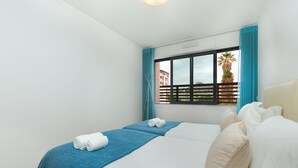 Een hotelkamer met een bed, een raam met gordijnen en een uitzicht op gebouwen en palmbomen.