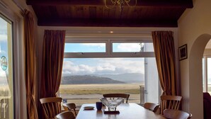 Een eetruimte met een tafel en stoelen, grote ramen met gordijnen en uitzicht op een landschap met bergen.