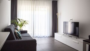 Una sala de estar moderna con un sofá gris, un televisor de pantalla plana sobre un mueble blanco y un ventanal con cortinas traslúcidas.