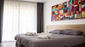 Una cama bien hecha con toallas enrolladas, un cuadro abstracto grande en la pared y cortinas translúcidas en la ventana.