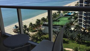 A balcony with two white cushioned chairs, overlooking a beach and a tennis court with palm trees. - Oceanfront renovated condo on the beach - amazing ocean view (Sunny Isles Beach)