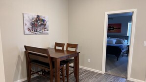 Un comedor con una mesa y sillas de madera, un cuadro en la pared y vistas a un dormitorio con cama y almohadas.