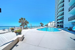Uma área de piscina com design curvo, cercada por um edifício branco com varandas, palmeiras e um céu azul límpido.