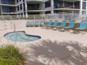 Zona de piscina con sillones de descanso y una piscina, rodeada por una valla blanca y un edificio con balcones.