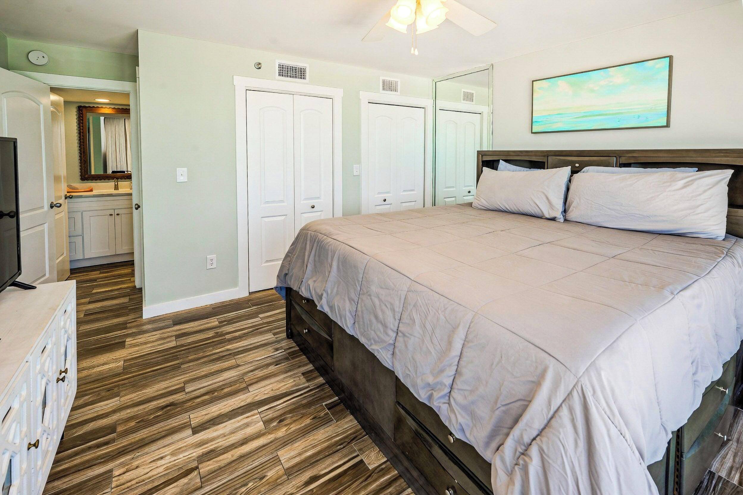 A bedroom with a large bed, wooden flooring, a ceiling fan, and a painting on the wall.