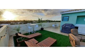 A rooftop terrace with wooden loungers, white tables, and chairs, a barbecue grill, and a view of the cityscape during sunset. - 5 pools, Amazing Rooftop Views , Beach (Vieques)