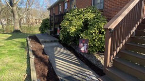 Un immeuble résidentiel à la façade en briques, doté d’un escalier en bois et d’un jardin agrémenté de paillis et d’une pancarte sur laquelle on peut lire : « J’aime mon chien. »