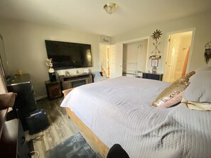 A bedroom with a large bed, a television, a dresser, and a door leading to another room.