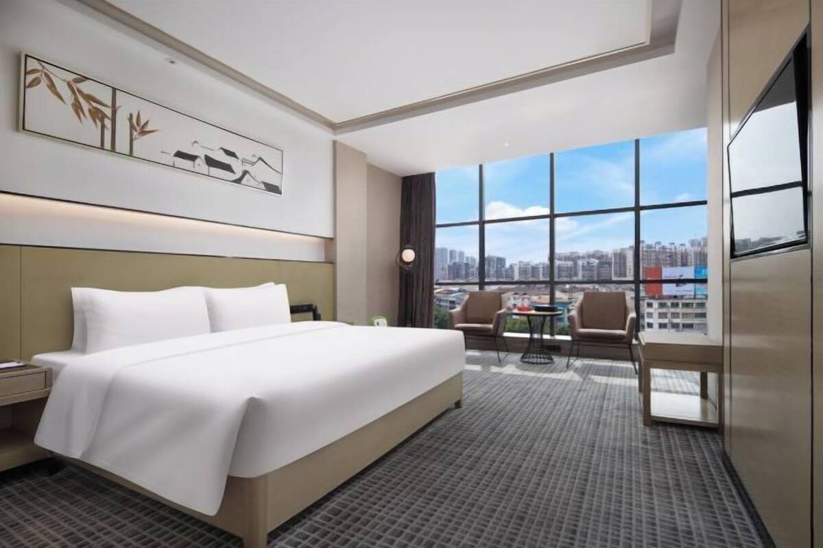 Habitación de hotel con una cama grande, un escritorio, una silla y vistas a la ciudad a través de amplias ventanas.