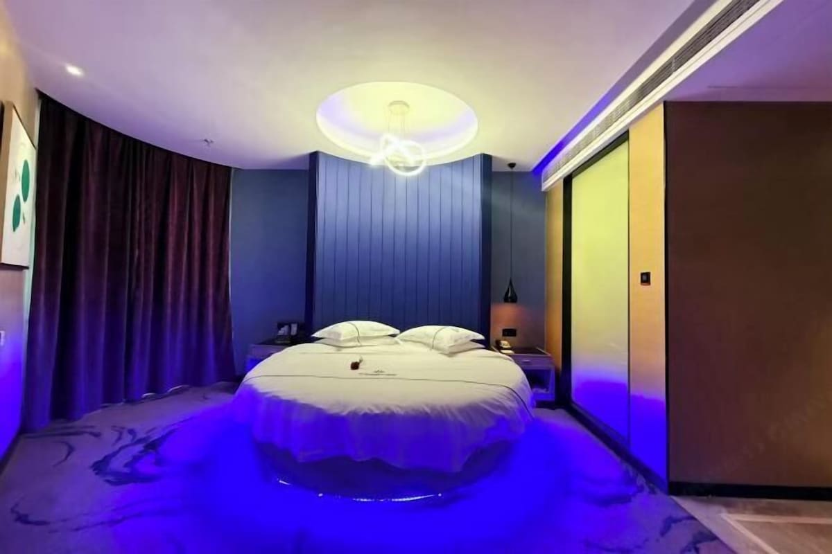A modern hotel room with a curved bed, a round ceiling light, and a view of the city.