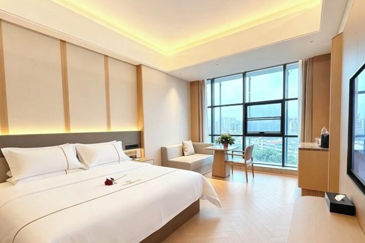A modern hotel room with a large bed, a seating area, and a view of the city through large windows.