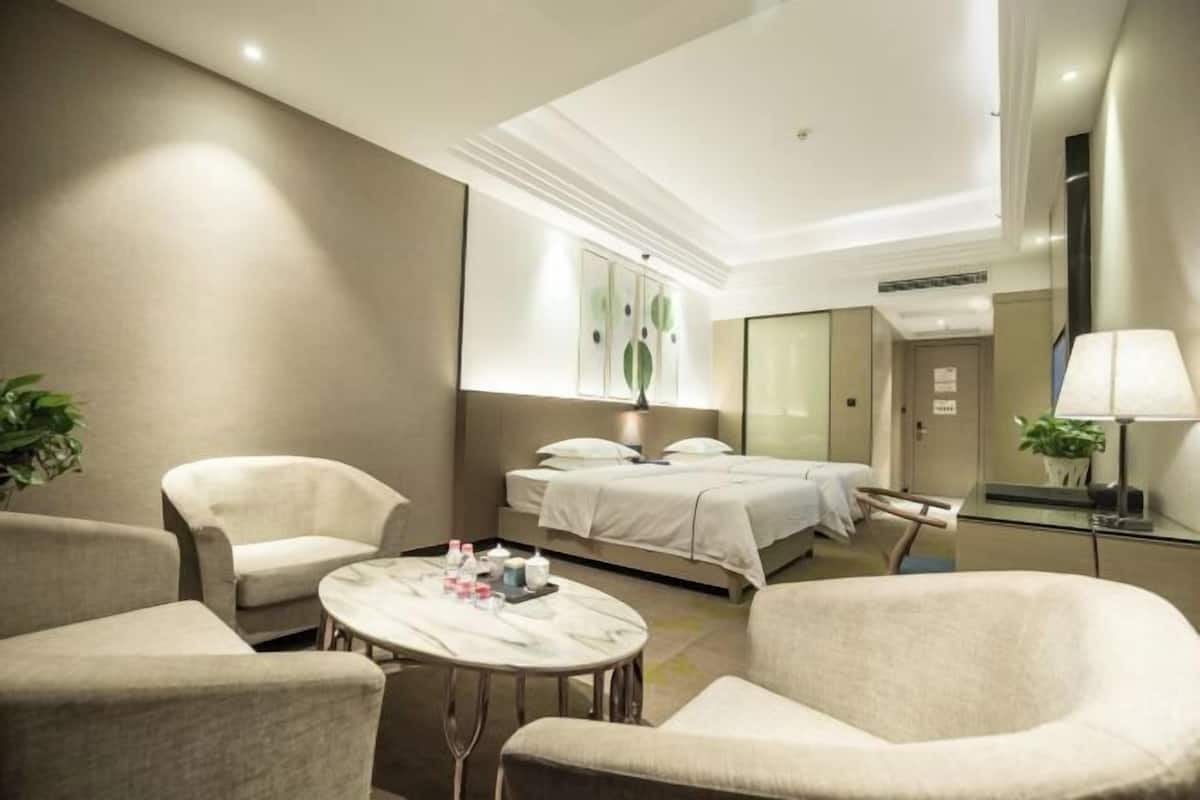 A hotel room with a bed, two armchairs, a round table with amenities, a mirror, and a plant.