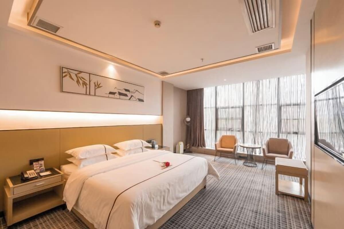 A hotel room with a large bed, a desk with a lamp, a chair, and a view of the outside through large windows.