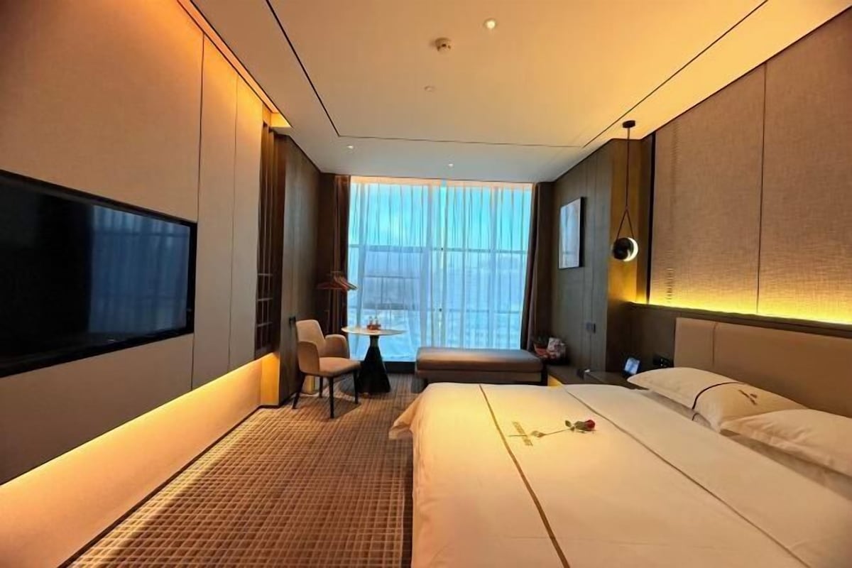 A hotel room with a large bed, a desk, a chair, a TV, and a window with curtains.