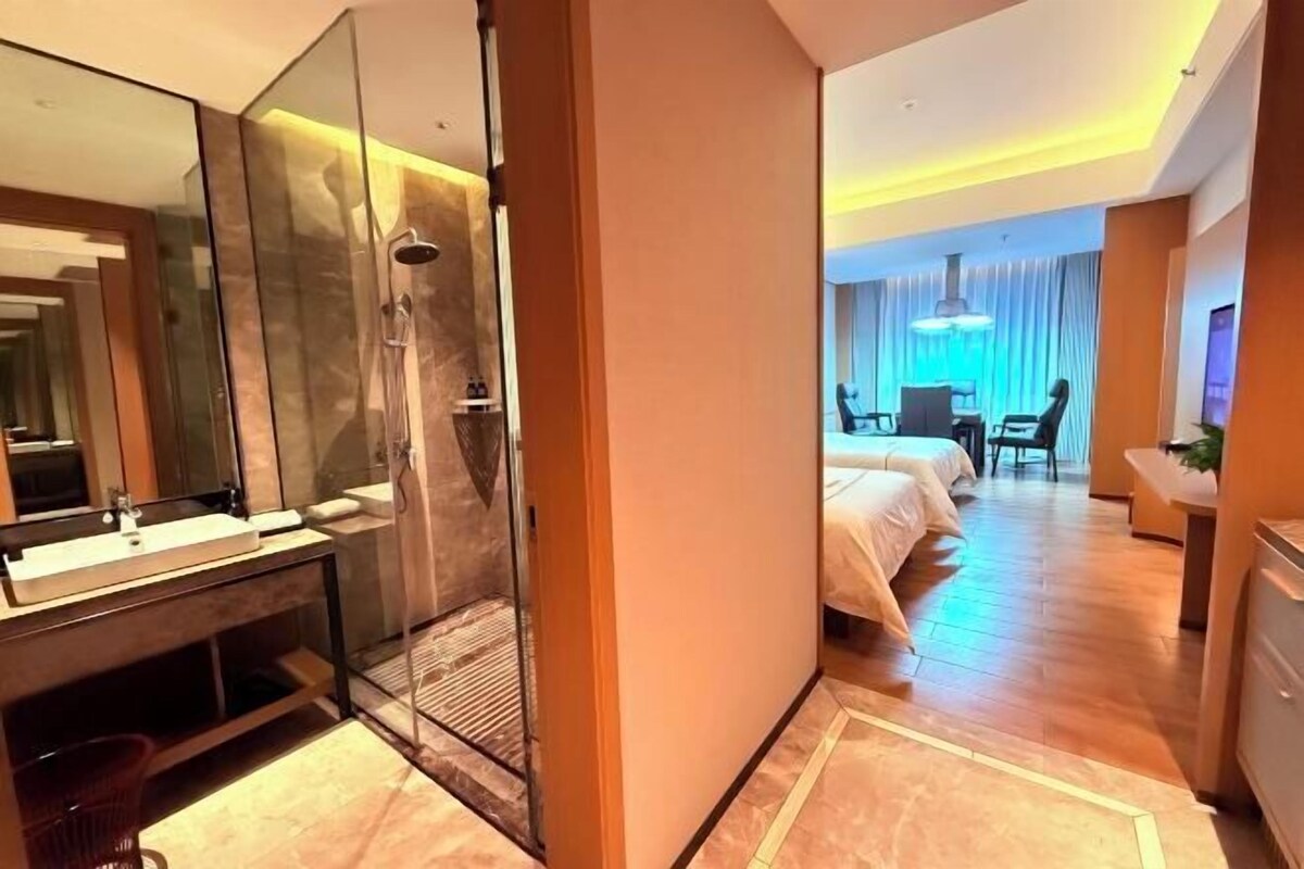 A hotel room with a large mirror, a shower, a bed, a desk, and a chair.