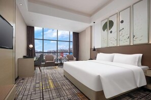 Habitación de hotel con una cama grande, un escritorio, una silla, un televisor y vistas a la ciudad a través de amplias ventanas.