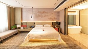 A modern hotel room with a large bed, a bedside table, a desk, a chair, a bench, a large window, a wall-mounted artwork, a bathroom with a bathtub, and a ceiling light. - Huarong Hongfa International Hotel (People's Square) (Huarong)
